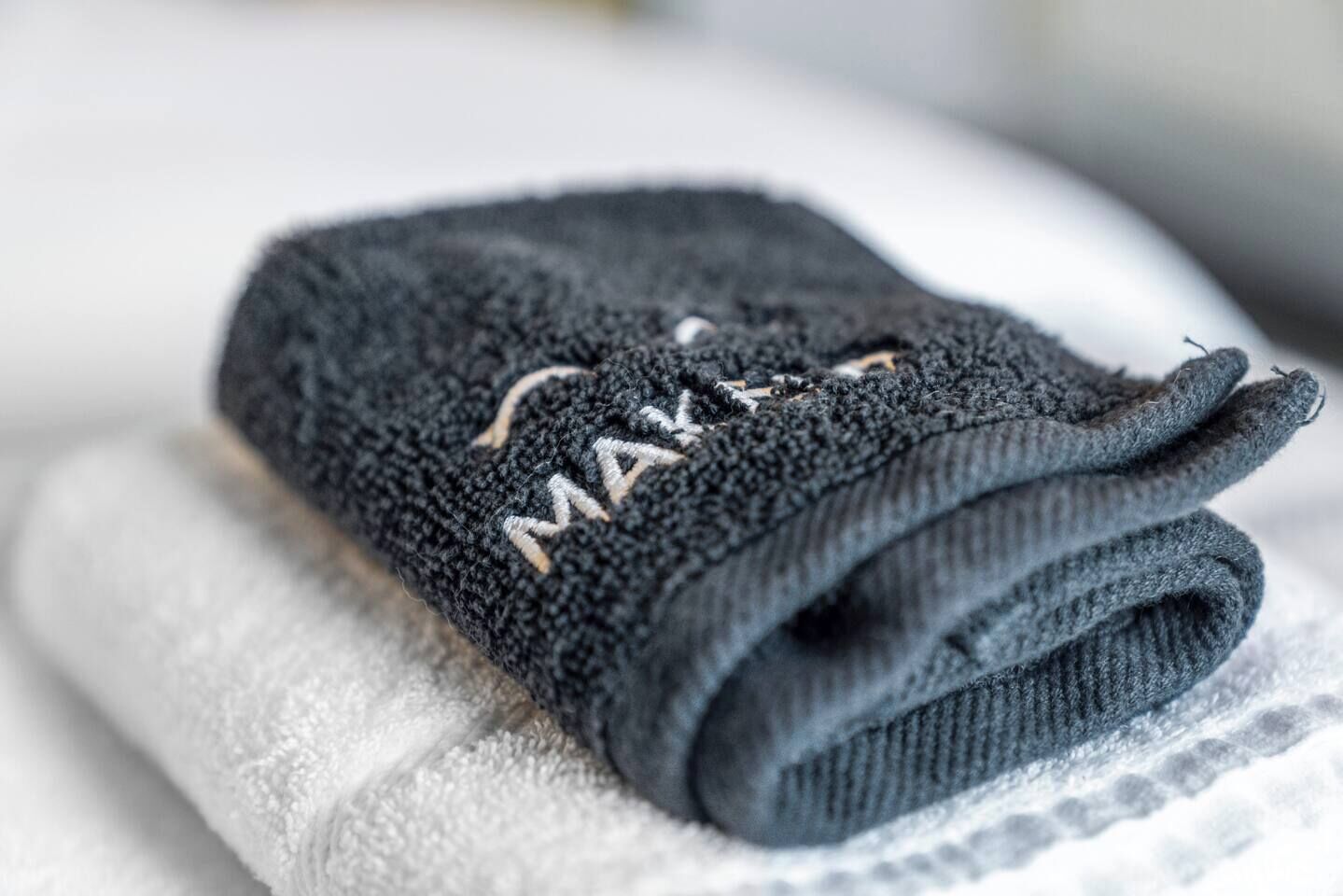 Une serviette pliée de couleur foncée, ornée d’une broderie bien visible « MA », est posée sur une serviette blanche.