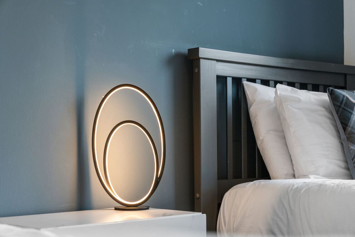 Een bed met witte beddengoed, een houten hoofdbord en een nachtkastje met een ronde lamp.