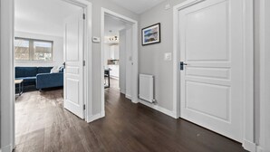 A hallway with white doors, a blue sofa, a kitchen in the background, and a framed picture on the wall. - Modern Stirling Apartment - Free Parking (Stirling)