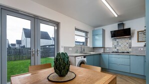 A modern kitchen with a washing machine, a dining table with a cactus centerpiece, and a view of a residential area through a glass door. - Modern 2-Bed Stirling House | Driveway Parking (Stirling)