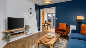 A modern living room with a TV, wooden coffee table, blue sofa, orange armchair, and a dining area in the background. - Modern 2-Bed Stirling House | Driveway Parking (Stirling)