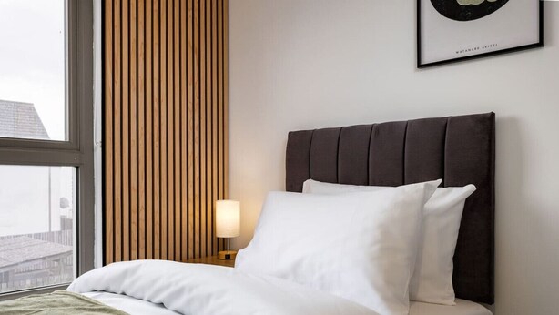 A hotel room with a bed, a nightstand, a lamp, a window, and a framed picture on the wall. - Modern 2-Bed Stirling House | Driveway Parking (Stirling)
