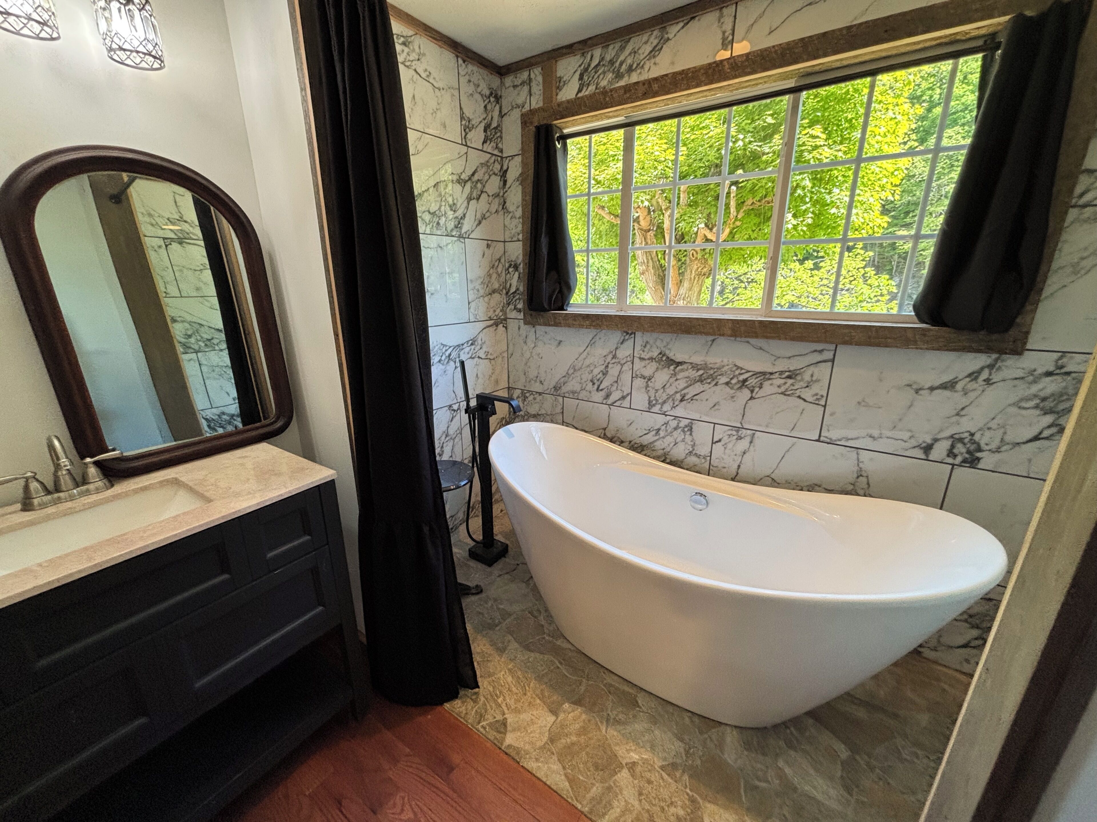 Un baño con bañera independiente, tocador con espejo y ventana con vista a vegetación.