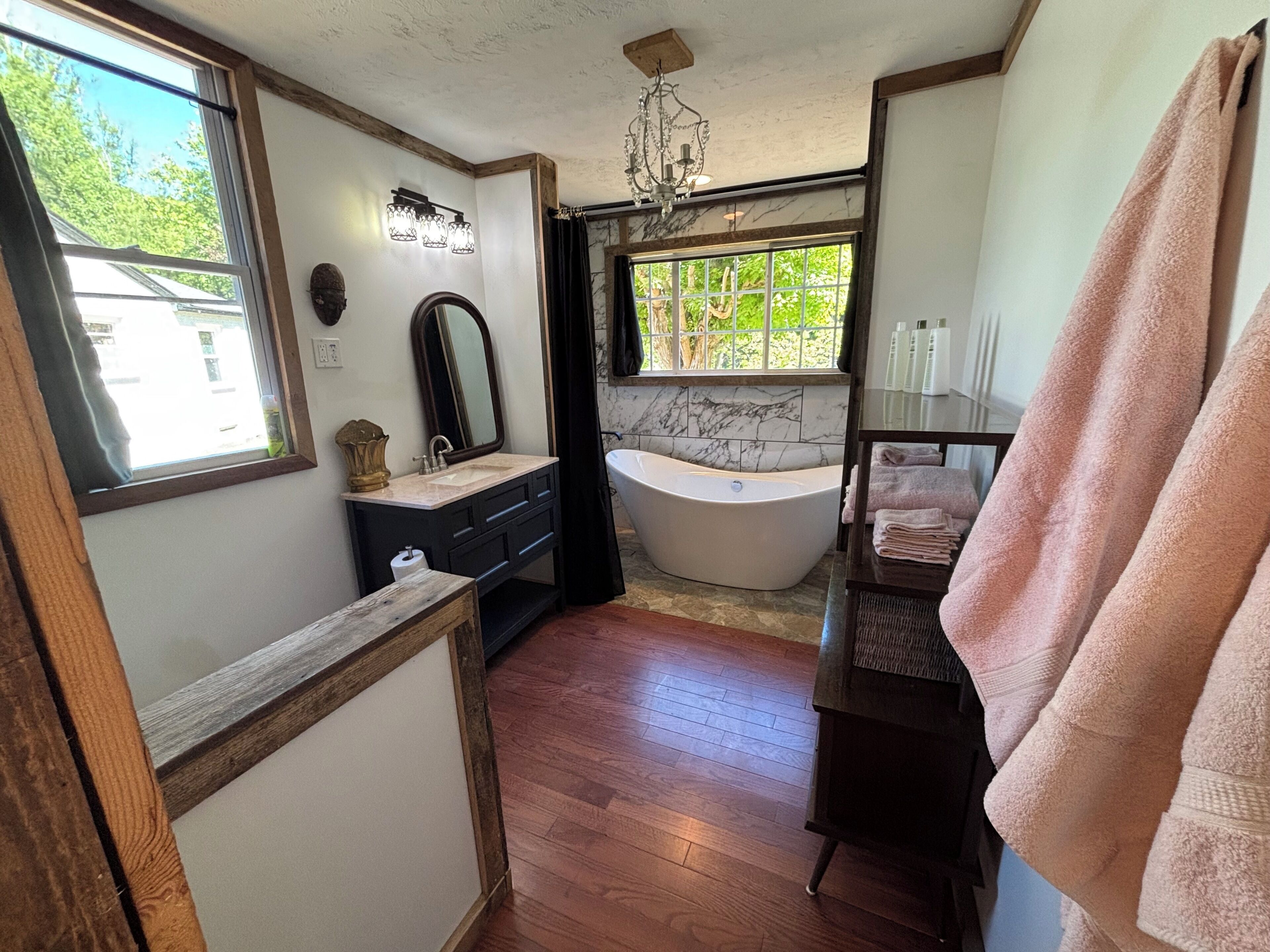 A bathroom with a bathtub, a vanity with a mirror, a chandelier, and a window with a view of greenery.