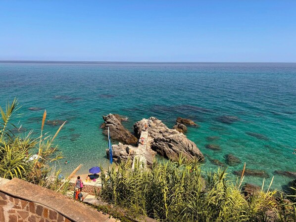 Blick auf die Küste mit klarem türkisblauem Wasser, Felsformationen und einer Steintreppe, die zum Strand hinabführt.