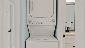Een voorzichtige kijk op een witte wasmachine en droger combinatie met bedieningspanelen, staand in een hal met lichtblauwe muren en een zichtbare badkamer met een wastafel en handdoekhouder.
