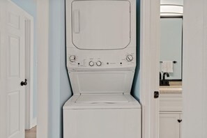 Eine Frontansicht einer weißen Waschmaschine und Trockner-Kombination mit Bedienfeldern, platziert in einem Flur mit hellblauen Wänden und einem sichtbaren Badezimmer mit Waschbecken und Handtuchhalter.