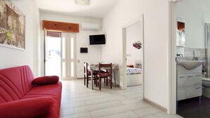 Ein Wohnzimmer mit rotem Sofa, einem Essbereich mit Tisch und Stühlen, einem an der Wand befestigten Fernseher und einer Küchenzeile mit Spüle und Schränken.