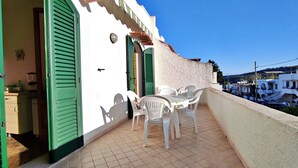 Un balcón con una mesa y sillas, una puerta con contraventanas verdes y vistas a un edificio blanco con pared texturizada.