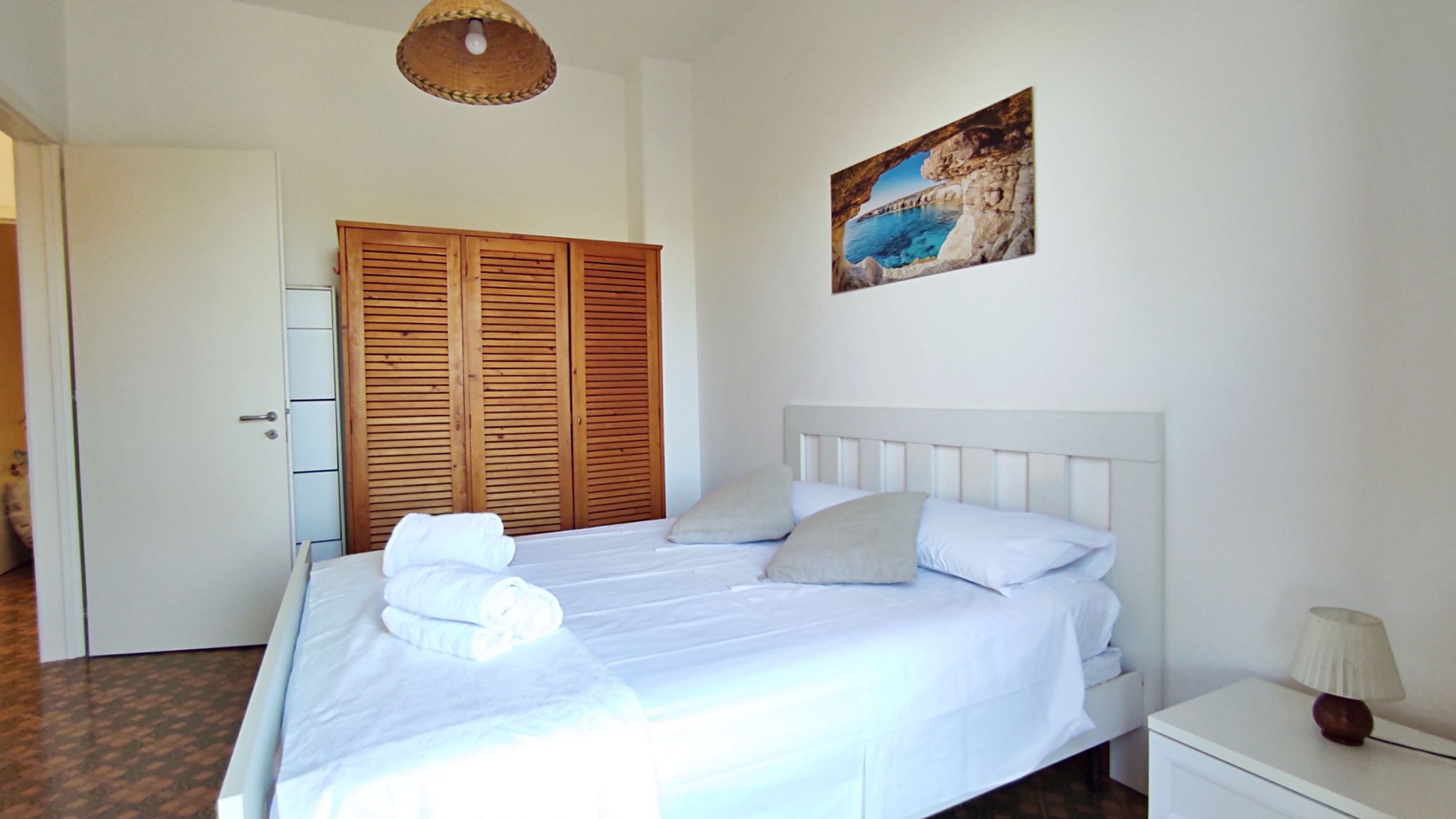 Um quarto com uma cama, mesa de cabeceira, abajur, guarda-roupa de madeira e um quadro na parede.