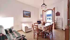 Una sala de estar con un sofá, una mesa de comedor con sillas, un cuadro en la pared y vistas a través de un ventanal abierto.