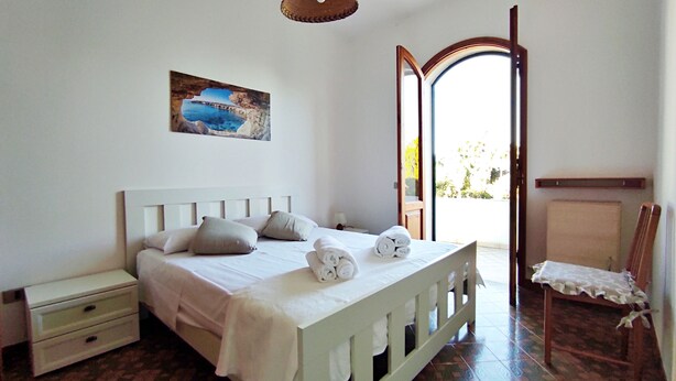 Un dormitorio con cama, mesita de noche, silla, un cuadro y una puerta abierta que da a un balcón.