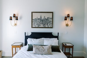 A bed with a dark headboard, white bedding, and multiple pillows. There are two bedside tables with lamps, a framed cityscape picture above the bed, and a small table with a remote control.
