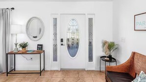 Une porte blanche avec un panneau de verre, un miroir rond, une petite table avec une lampe, un canapé en osier et un tableau encadré accroché au mur.