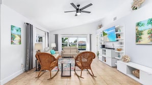 Un salon avec un ventilateur de plafond, deux fauteuils en osier, un canapé beige, une table basse et un téléviseur posé sur une étagère blanche.