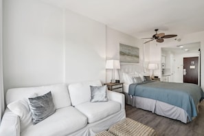 Ein Hotelzimmer mit einer weißen Couch, einem Bett mit blauer Bettdecke, einem Deckenventilator und einem Bild an der Wand.