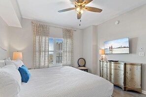 A hotel room with a large bed, a ceiling fan, a TV, and a view of a building through the window. - Majestic Sun 1108b - Wave Whisper (Miramar Beach)