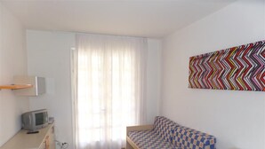 Ein kleiner, heller Raum mit einem Bett, einem Schreibtisch, einem Stuhl, einem Fernseher und einem Fenster mit Vorhängen.