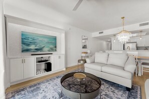 A modern living room with a large flat-screen TV, a white sofa, a round coffee table, and a chandelier. - Majestic Sun 312b - 3 Tortugas (Miramar Beach)