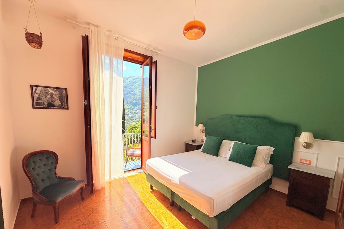 A bedroom with a bed, a chair, a nightstand, a lamp, a picture on the wall, and a view of the outdoors.