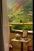 A balcony with a table, chairs, a potted plant, and a scenic view of a valley with buildings.