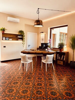 Un comedor con una mesa de madera, dos sillas blancas, una lámpara colgante, un televisor y una estantería con plantas.
