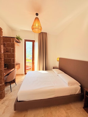 A bedroom with a large bed, a desk, a chair, a hanging light fixture, and a view through a door. - Putechella - Retro Rooms & Villas (Tramonti)