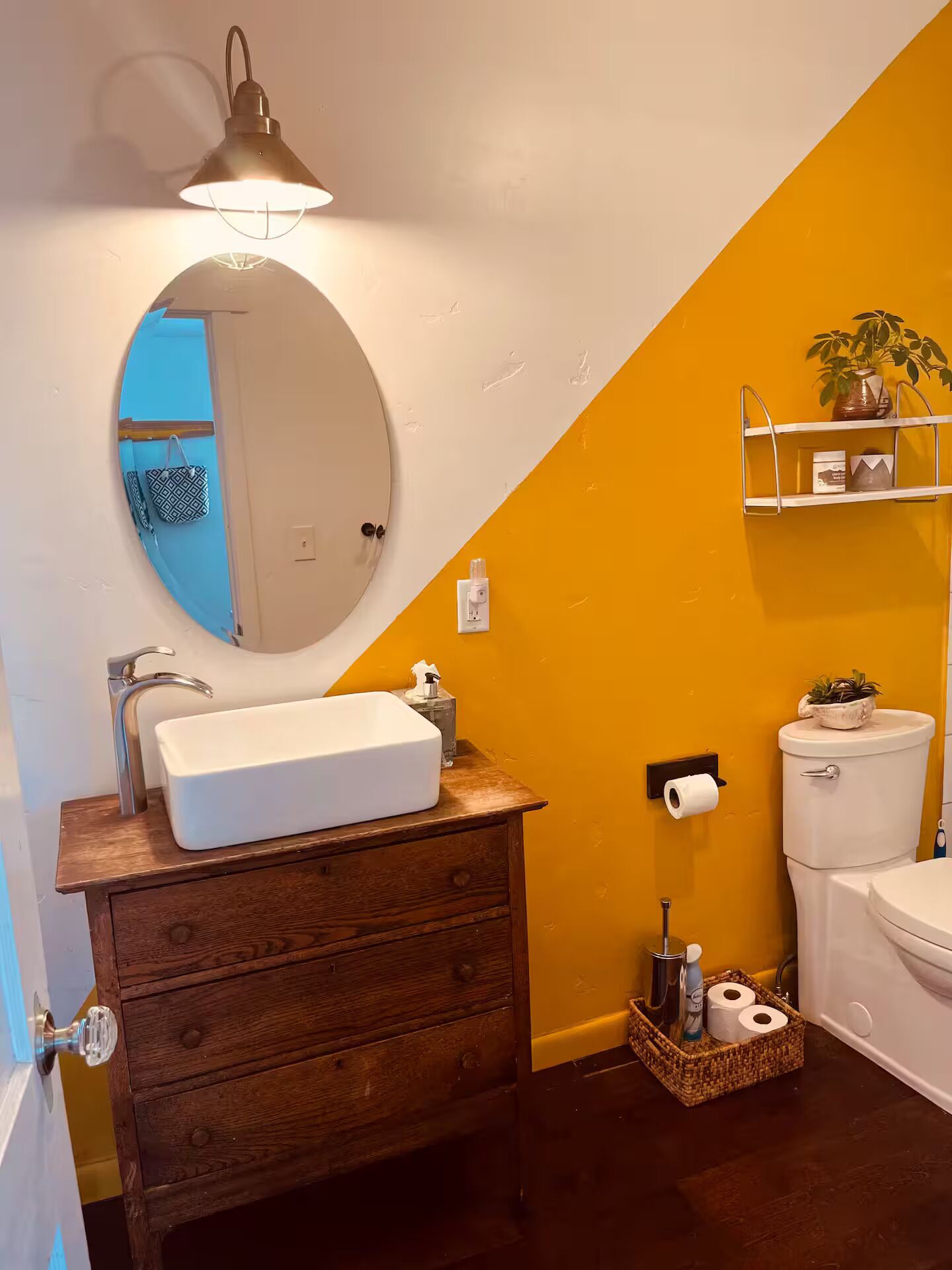 Une salle de bain avec un lavabo blanc, un meuble-lavabo en bois, un miroir rond, un mur d’accent jaune, des toilettes et une petite étagère avec une plante.