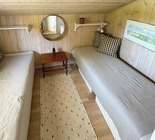 A cozy cabin interior with two single beds, a small table, a round mirror, and a painting on the wall. - Holiday home in scenic surroundings in Lønstrup (Hjørring)