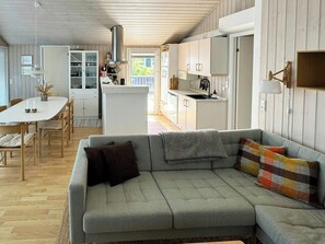 Un saló modern amb un sofà seccional gris, una taula de dinar amb caires, una zona de cuina amb armaris blancs i un sostre de fusta.