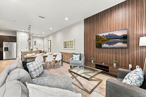 A modern living room with a large flat-screen TV mounted on a wooden wall, a dining area with a kitchen in the background, and a seating area with a glass coffee table. - Family Retreat with 4Bed suites & Private Jacuzzi (Four Corners)