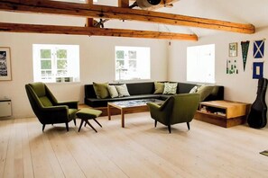 A cozy living room with a sofa, two armchairs, a coffee table, and a guitar. There are windows with blinds, wooden beams on the ceiling, and a wall with framed pictures and a flag.