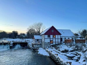 A traditional building with a red roof and white walls, situated by a river with a stone bridge.