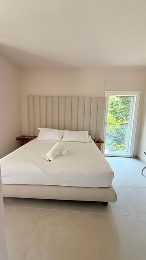 Una cama grande con cabecera, ropa de cama blanca y vistas a la vegetación a través de una ventana.