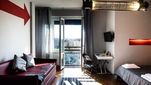 Hotellihuone, jossa on punainen sohva, sänky, työpöytä ja ikkunasta avautuva näkymä kaupunkiin.