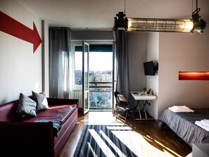 A hotel room with a red sofa, a bed, a desk, and a view of the city through the window. - CARLOTTA STATION SUITE (Bologna)