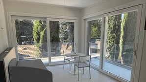 Ein modernes Zimmer mit einer Couch, einem kleinen runden Tisch und einem Stuhl, große Schiebeglasfenster zu einem Außenbereich mit Blick auf Bäume und eine Backsteinmauer.