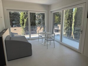 A modern room with a sofa, a small round table, and a chair, large sliding glass doors leading to an outdoor area with a view of trees and a brick wall. - Superior with Balcony (Bologna)