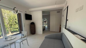 Ein modernes Wohnzimmer mit grauem Sofa, einem Esstisch mit Stühlen, einem an der Wand montierten Fernseher und einem großen Fenster mit Blick auf Bäume.