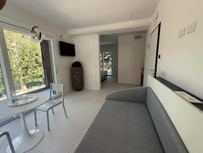 A modern living room with a grey sofa, a dining table with chairs, a TV mounted on the wall, and a large window with a view of trees. - Superior with Balcony (Bologna)