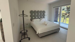 Ein Schlafzimmer mit einem Bett, einem Nachttisch, einem runden, an der Wand befestigten Kopfteil, einem Kleiderständer und einem großen Fenster mit Blick auf Bäume.