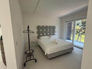 A bedroom with a bed, a nightstand, a wall-mounted circular headboard, a clothing rack, and a large window with a view of trees. - Superior with Balcony (Bologna)