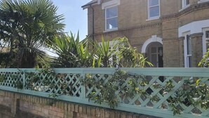 A brick house with a white window frame, a green fence, and a palm tree in the foreground. - Clarence House Holidays (Shanklin)