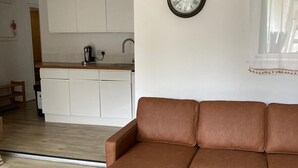 A living room with a brown sectional sofa, a white kitchen in the background, and a wall clock. - Clarence House Holidays (Shanklin)