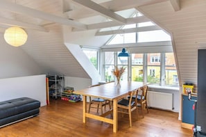 Ett rum i loftstil med ett matbord, stolar, en svart soffa, ett element, ett fönster med utsikt över byggnader och en hängande lampa.