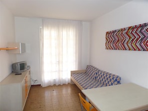 Ein kleiner, heller Raum mit einem Bett, einem Schreibtisch, einem Stuhl, einem Fernseher und einem Fenster mit Vorhängen.