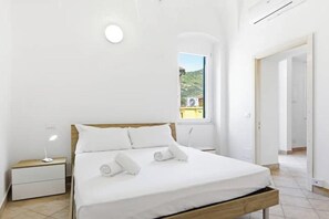 Ein modernes Schlafzimmer mit einem Holzbettgestell, weißer Bettwäsche, einem Nachttisch mit Lampe und einem Fenster mit Blick auf eine grüne Landschaft.