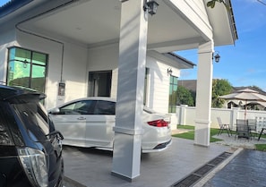 En lukket carport med en hvid bil parkeret indeni, en sort bil parkeret udenfor, og en patio med udendørs møbler.