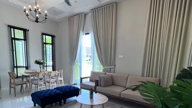 A modern living room with a sofa, ottoman, round coffee table, and a chandelier. There are two windows with blinds, a dining area with chairs, and a potted plant.