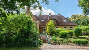 Une maison traditionnelle avec un toit en tuiles, un escalier en pierre et une végétation luxuriante qui l’entoure.